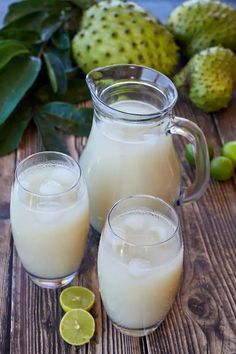 two glasses filled with white liquid next to some limes on a wooden table and green leaves