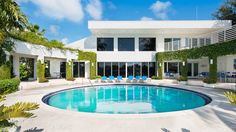 a large swimming pool in front of a white house with palm trees and lawn chairs