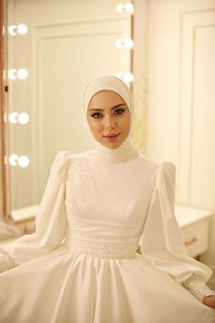 a woman in a white dress sitting on a bed with a mirror and lights behind her