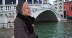 an older woman standing on the side of a river next to a bridge and buildings