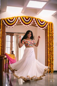 a woman in a white sari dancing under an arch decorated with flowers and garlands