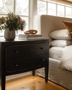 a bed with white sheets and pillows next to a nightstand in front of a window