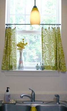 a kitchen sink under a window with green curtains and a yellow light hanging above it