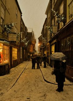 people walking down an alley way in the snow with umbrellas and lights on buildings