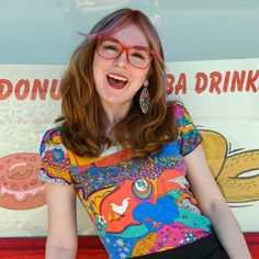 Closeup of happy model in brightly colored shirt in front of a bakery Multicolor Tops With Funny Print For Spring, Fun Multicolor Printed T-shirt, Retro Green Printed T-shirt, Colorful Printed Short Sleeve Top, Fun Multicolor Print T-shirt For Spring, Fun Multicolor Tops With Graphic Print, Fitted Summer Tops With Retro Print, Funny Print Multicolor Short Sleeve Tops, Spring Multicolor Tops With Unique Print