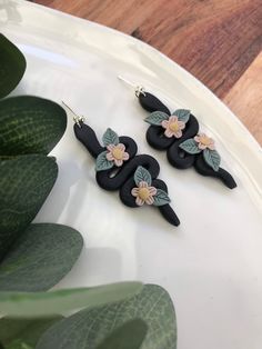 three black and pink flower earrings on a white plate next to green leaves, with a plant in the background