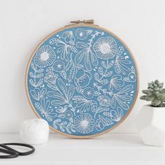 a blue and white embroidery pattern on a wooden frame next to a potted plant