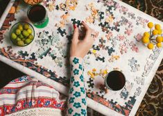 a person is working on some puzzles with cups of coffee and bowls of fruit