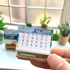 a miniature calendar is held in front of a desk with succulents and potted plants