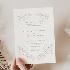 a person holding up a wedding card in front of flowers and greenery on a table