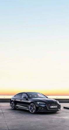 a black car parked on top of a cement parking lot next to the ocean at sunset