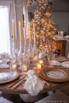 a dining room table is set for christmas with candles and plates on it, along with a lit tree in the background