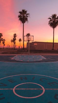 an outdoor basketball court with palm trees and the sun setting in the sky behind it