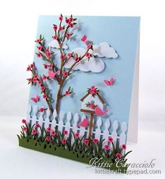 a handmade card with pink flowers and a bird house in the grass next to a white picket fence