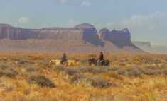 two people riding horses through the desert with mountains in the background