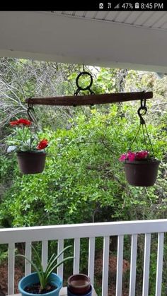 two potted plants hanging from a wooden beam on a porch with white railing and trees in the background