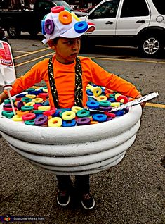 a young boy in an orange shirt and hat holding a large white bowl filled with doughnuts