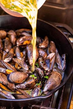 a pan filled with mussels being cooked on an open fire grill by someone pouring sauce over them