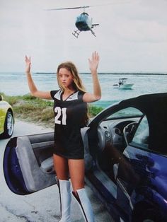 a beautiful young lady standing in front of a car next to a helicopter flying over the ocean