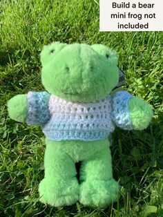 a green teddy bear wearing a blue and white crochet shirt sitting in the grass