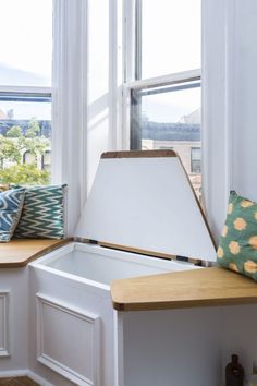 a white chest sitting in front of a window next to a wooden table with pillows on it