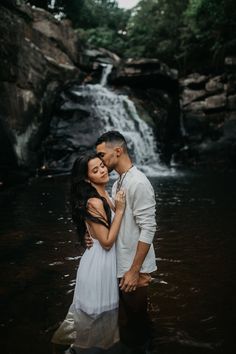 a man and woman standing in the water next to a waterfall with their arms around each other