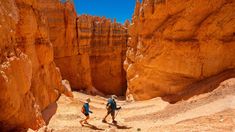 two people hiking in the desert with large rocks on either side and one person walking through between them