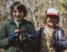 two young boys are standing in the woods with their cameras and smiling at the camera