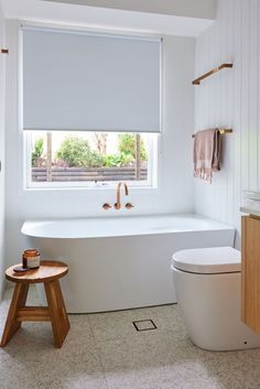 a white bath tub sitting under a window next to a toilet and wooden stool in a bathroom