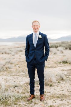 a man in a suit and tie standing in the middle of a desert field with his hands in his pockets