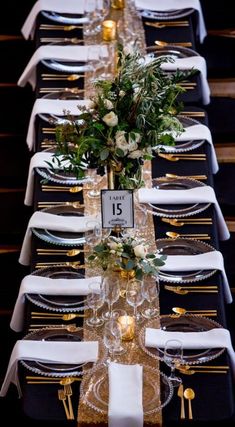 a long table is set with black and white linens, gold place settings, greenery and candles