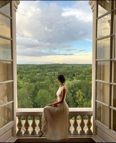 a woman in a long dress sitting on a balcony looking out the window at trees