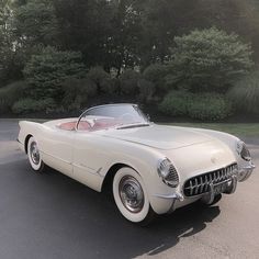 an old fashioned white car parked in a parking lot next to some trees and bushes