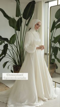 a woman in a white wedding dress standing next to a large potted plant and mirror