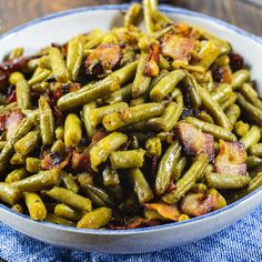 a bowl filled with green beans and bacon on top of a blue cloth next to a wooden table