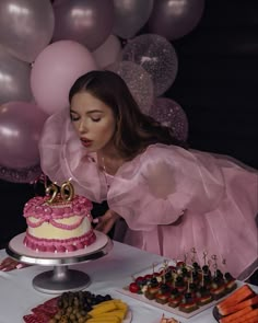 a woman in a pink dress blowing out candles on a cake with balloons behind her