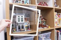 a person holding up a toy house in front of a book shelf filled with toys