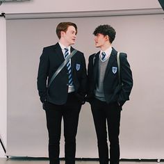 two young men standing next to each other in front of a white wall wearing suits and ties