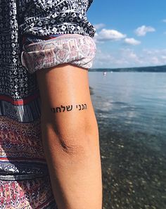 a woman's arm with the word hebrew tattooed on it, in front of a body of water