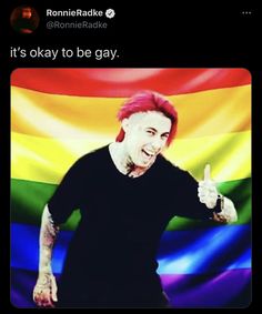 a man with pink hair and piercings giving the thumbs up in front of a rainbow flag