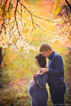 a pregnant couple standing under a tree in the fall