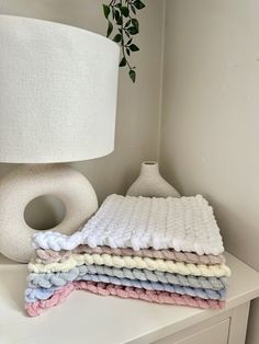 a stack of blankets sitting on top of a white table next to a lamp and potted plant