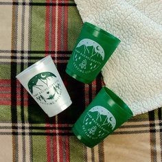 three green cups sitting on top of a plaid blanket next to a white and black towel