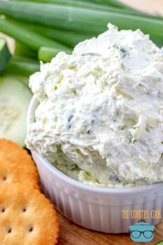 a white bowl filled with cream cheese surrounded by crackers and cucumbers