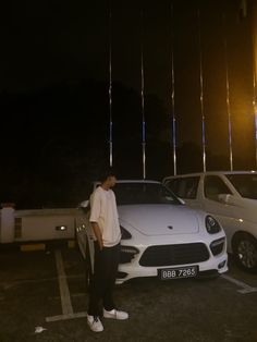 a man standing next to a white car in a parking lot with lights hanging from the ceiling