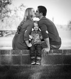 a man and woman kiss their son as they sit on a wall in the grass