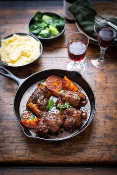 some meat and vegetables in a pan on a wooden table next to glasses of wine