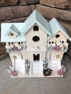 a white bird house with flowers and birds on the roof is sitting in front of a stone wall