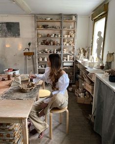 a woman sitting at a table in a room with pottery on the walls and shelves