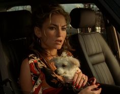 a woman sitting in the back seat of a car holding a small white dog and looking at the camera